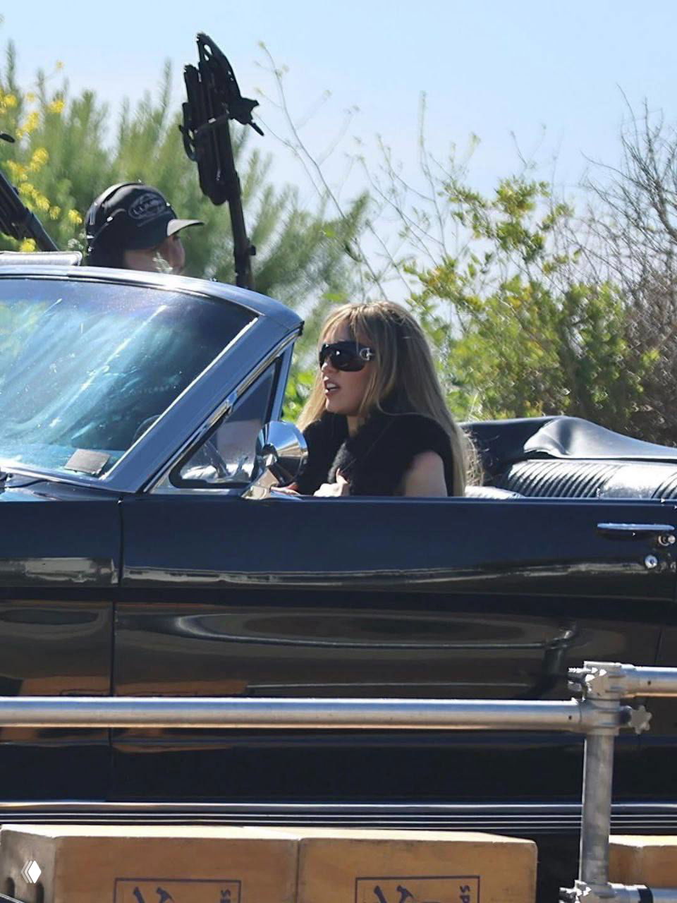 Крупный план женщины за рулём черного кабриолета на внешней локации в Малибу; видны осветительное оборудование и бум-микрофон.