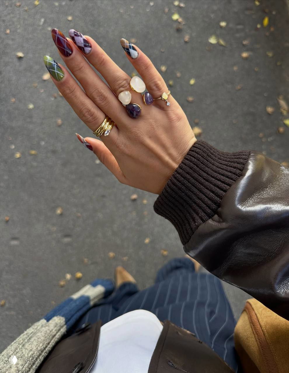 Рука с маникюром аргайл на фоне асфальта и осыпавшихся листьев; видны украшения, чёрная кожаная куртка и элементы одежды.