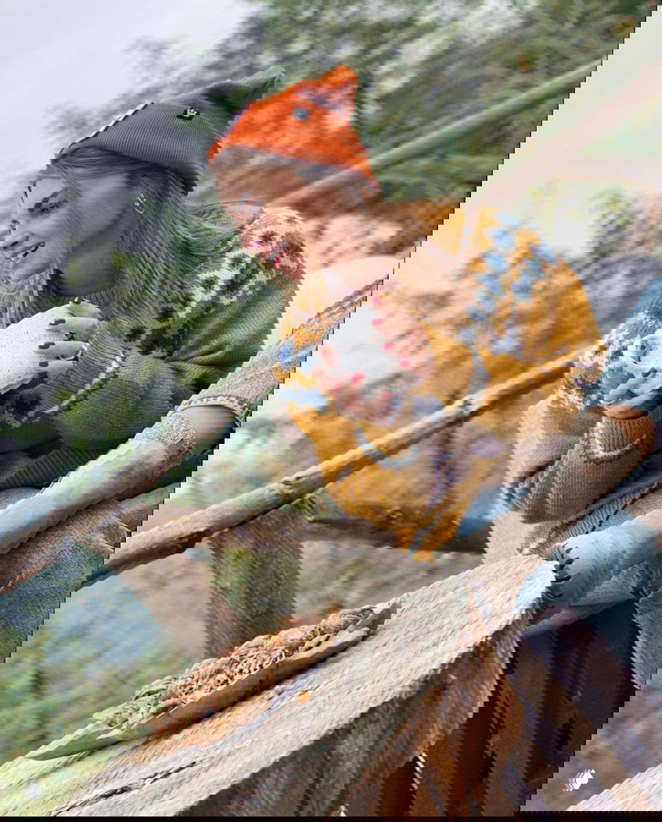 Кадр на открытой веранде в природной обстановке: женщина в тёплом свитере и оранжевой шапке держит кружку, на столе тарелка с орехами и лесной фон.