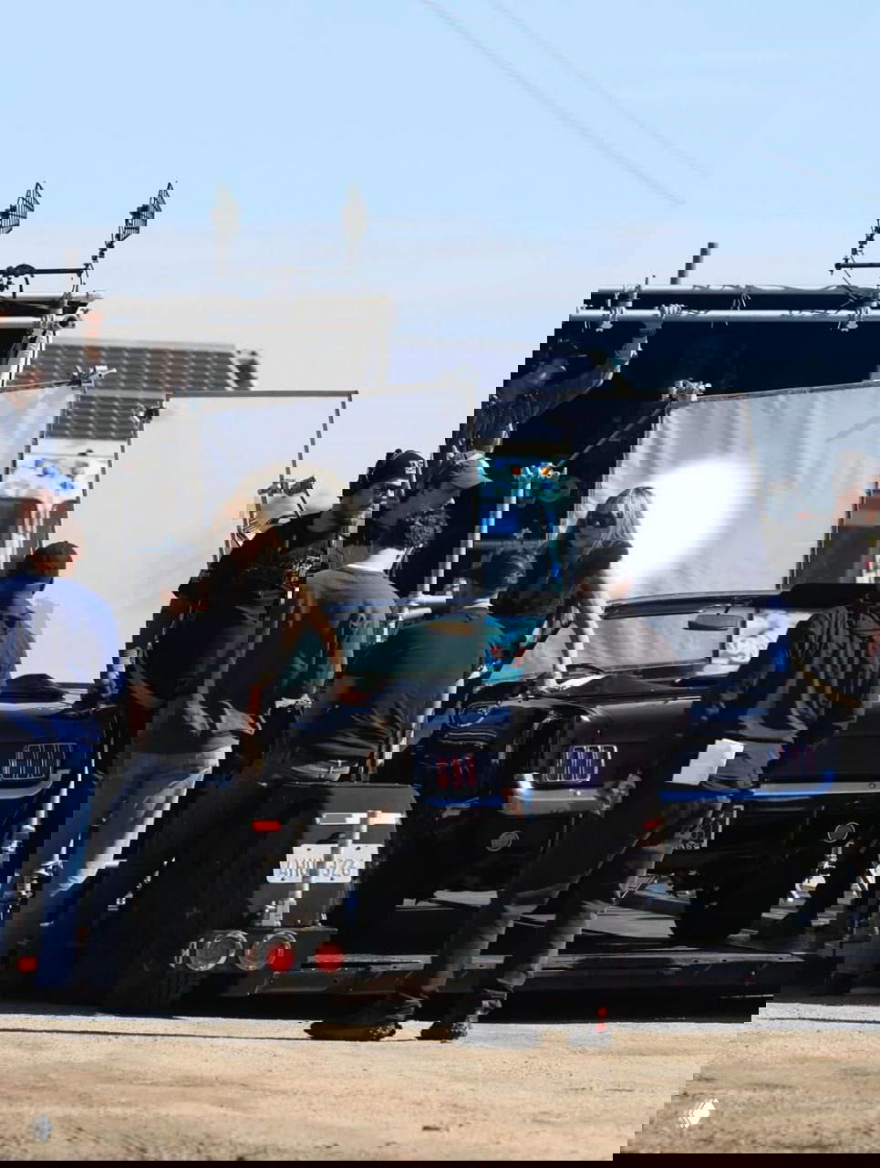 Общий план съёмочной площадки в Малибу: черный кабриолет, женщина рядом с машиной и несколько членов команды с техникой и реквизитом.