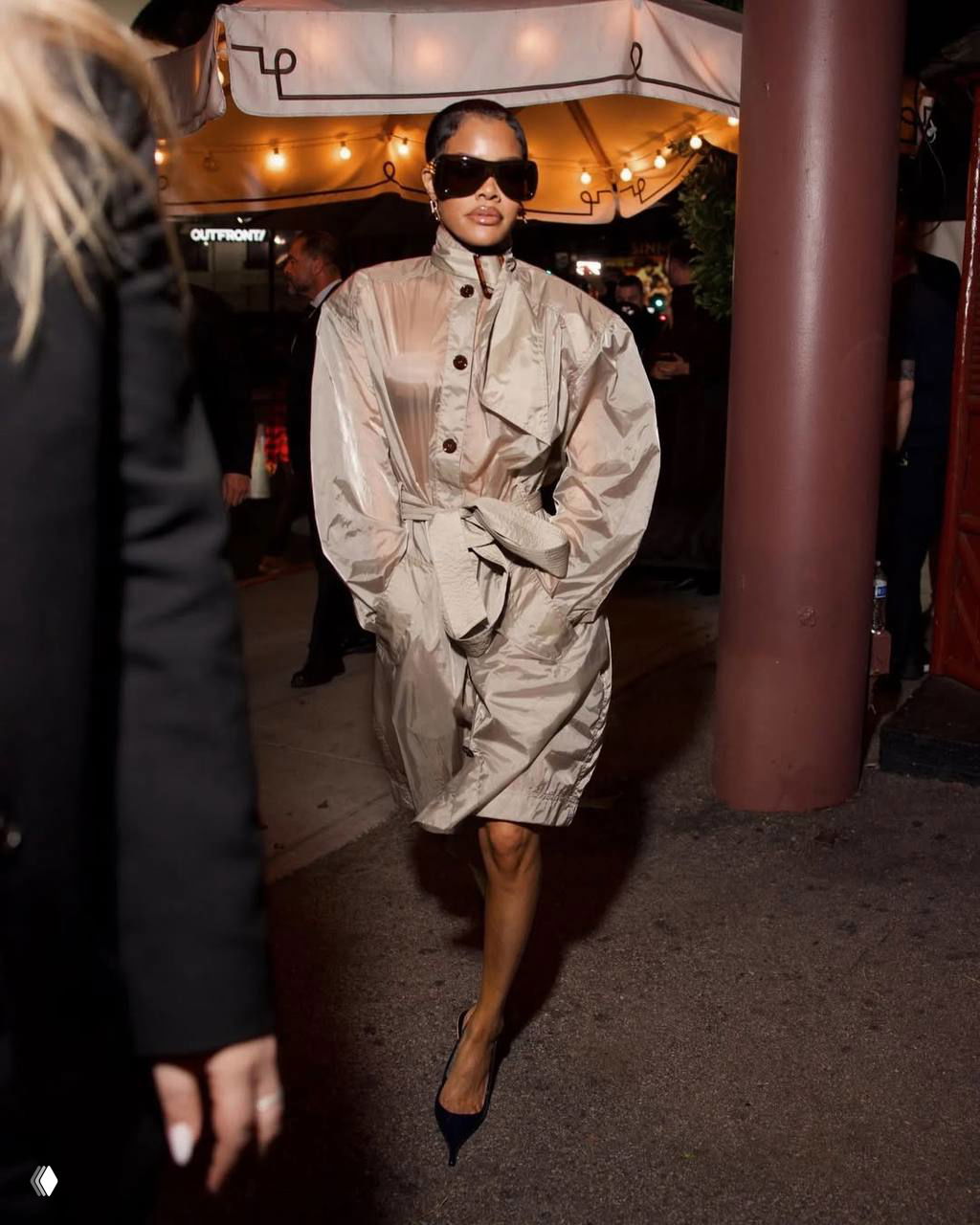 Гостья в просторном лёгком пальто‑плаще идёт по тротуару после афтепати «Грэмми», крупные солнцезащитные очки и вечерняя атмосфера.