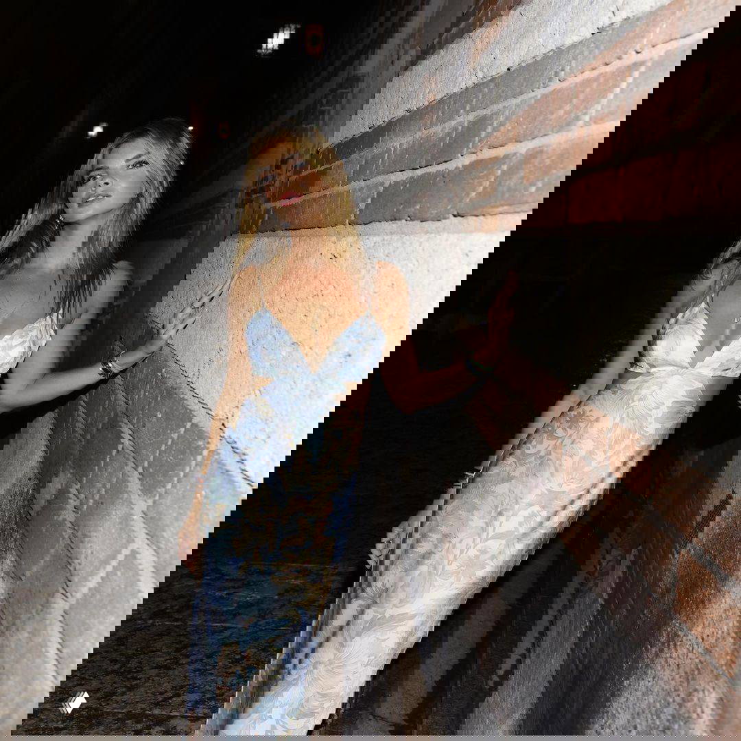 Ночной портрет женщины в длинном лёгком платье, она опирается на кирпичную стену узкой улочки; тёплое освещение создаёт атмосферу вечера.