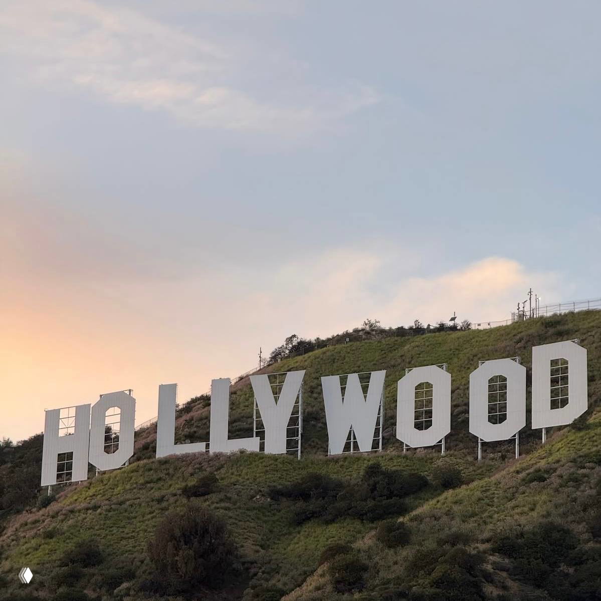 Знаменитая вывеска HOLLYWOOD на холме при нежном закатном освещении, пастельное небо и зелёные склоны — ассоциация с Лос‑Анджелесом.