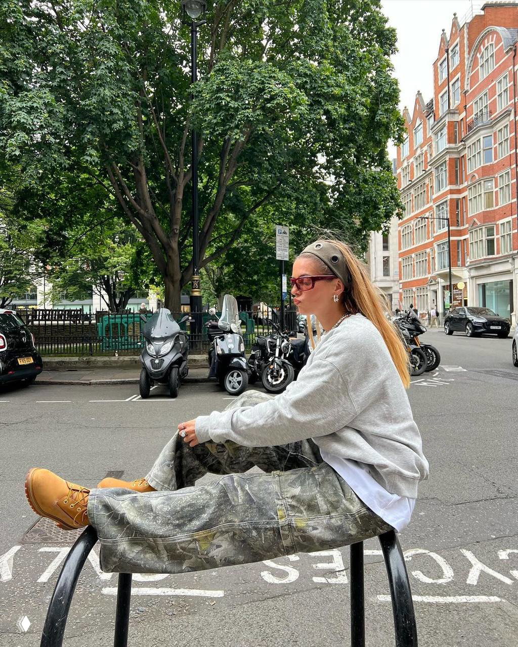 Модель в серой толстовке и свободных камуфляжных брюках сидит на урбанистической скамье — образ с рабочими ботинками и повседневной эстетикой.