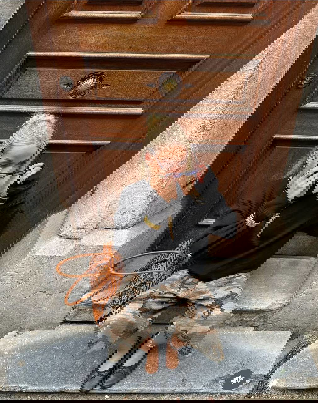 Блондинка в чёрном свитере и камуфляжных широких брюках сидит у массивной деревянной двери — сочетание грубой фактуры и лаконичного верха.