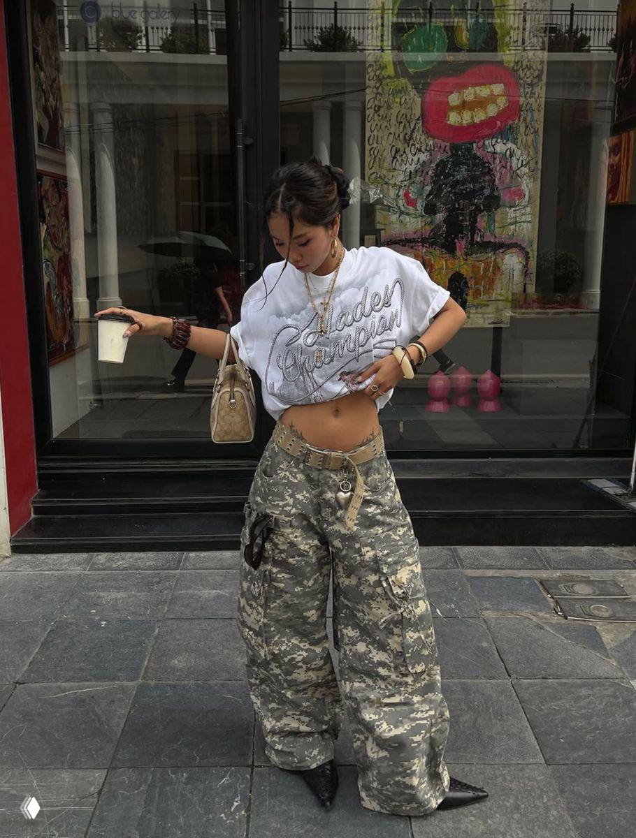 Девушка в белой укороченной футболке и камуфляжных шароварах идёт по городской брусчатке с бумажным стаканом в руках — лёгкий летний вариант с милитари-принтом.