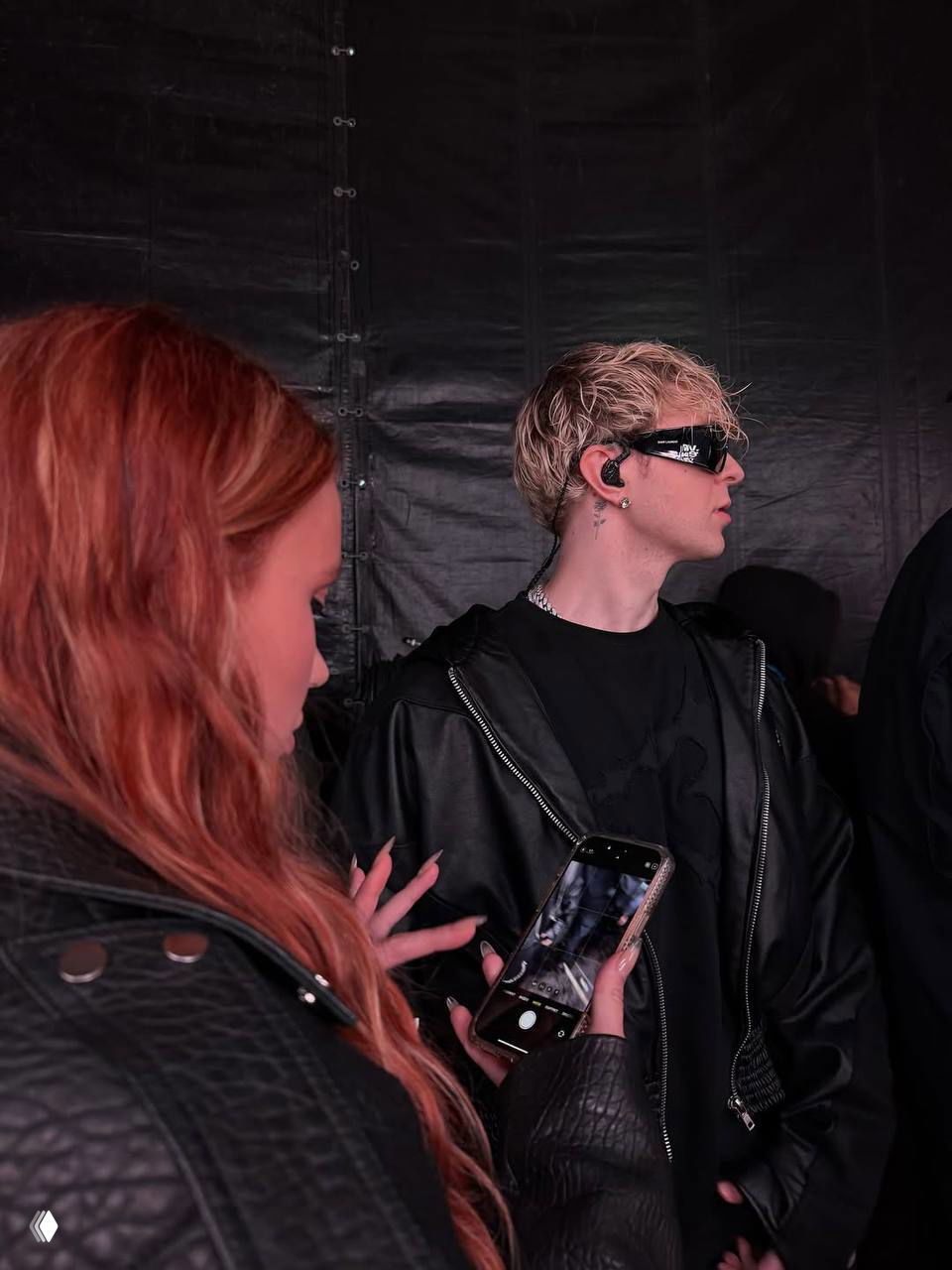 За кулисами: женщина с рыжими волосами держит смартфон, рядом мужчина в тёмной одежде и наушниках; кадр в контексте музыкального или фестивального события.