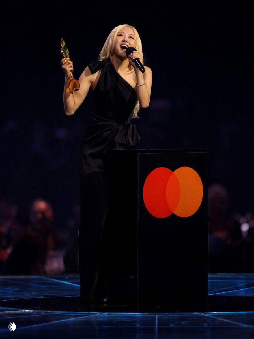 Полный рост Розэ на сцене BRIT Awards: чёрное платье, статуэтка в руке, за ней подиум с логотипом Mastercard и световое оформление сцены.