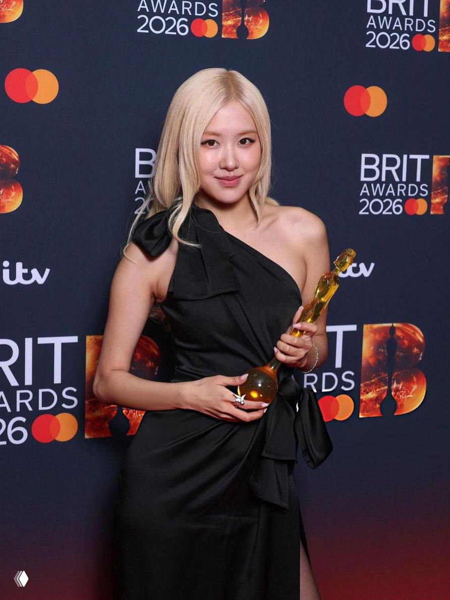 Портретное фото Розэ на красной дорожке BRIT Awards 2026: чёрное асимметричное платье, в руках прозрачная золотистая статуэтка.