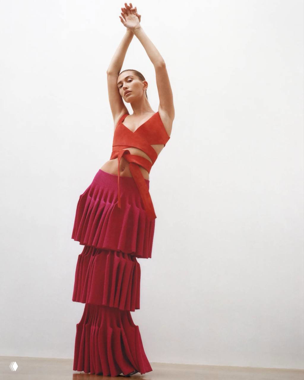 Хейли Бибер в длинной многослойной красной юбке и топе Alaïa, полностраничный кадр на нейтральном фоне, поза с поднятыми руками.