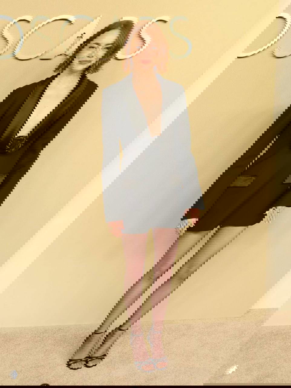 Фотография в полный рост: женщина в коротком чёрном мини‑платье с выразительным декольте и тонкими босоножками на бежевом фоне.