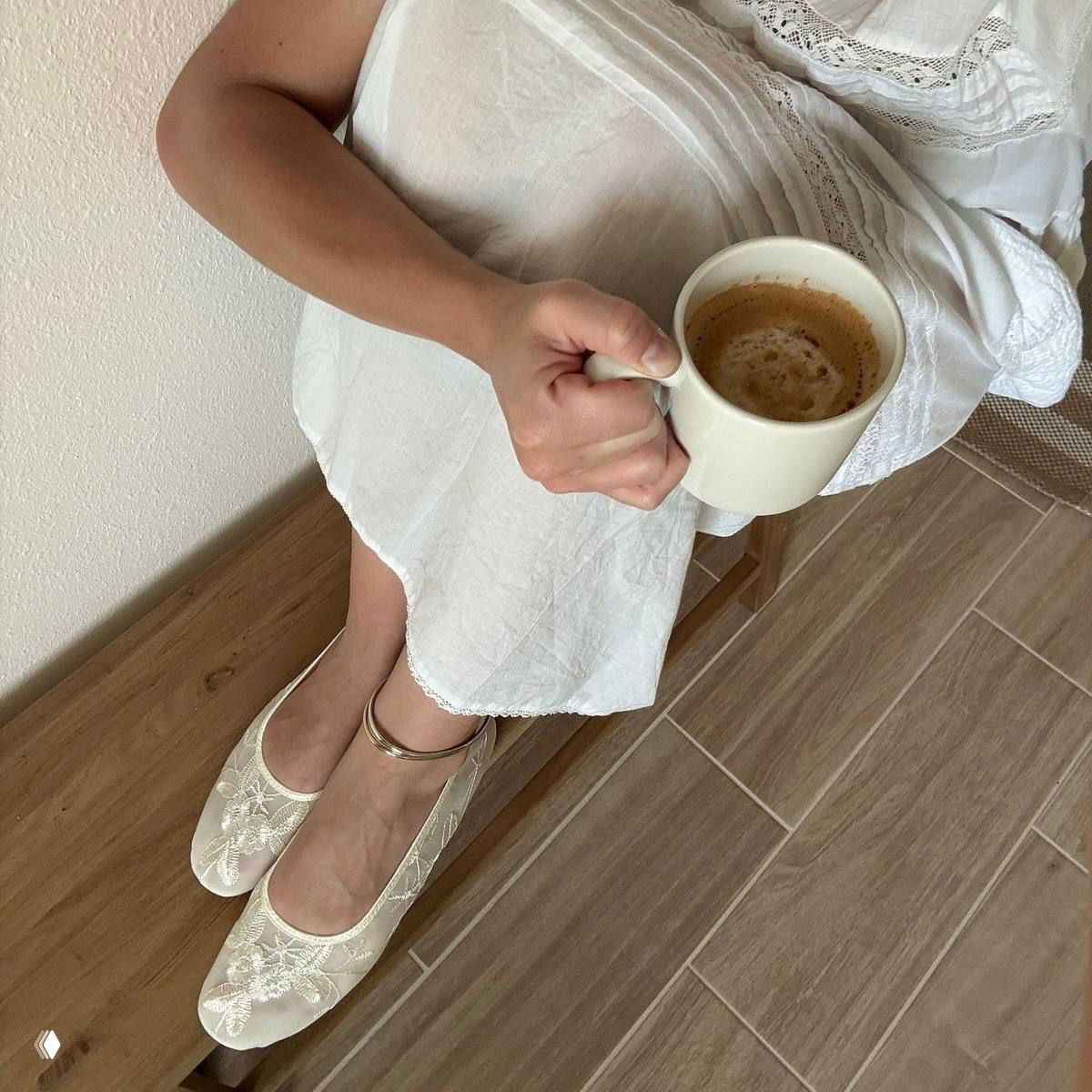 Девушка в лёгком белом платье держит чашку кофе, кружевные балетки и деревянный пол — нежная бытовая сцена в ретро-стиле.