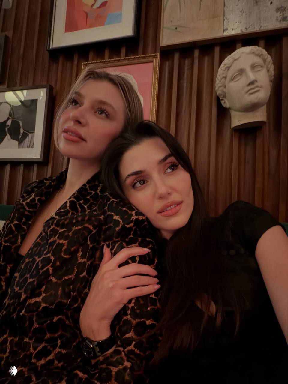 Две молодые женщины на селфи в тёплом интерьере кафе или ресторана: мягкое освещение, дружеская поза и интерьер с картинами на стене.