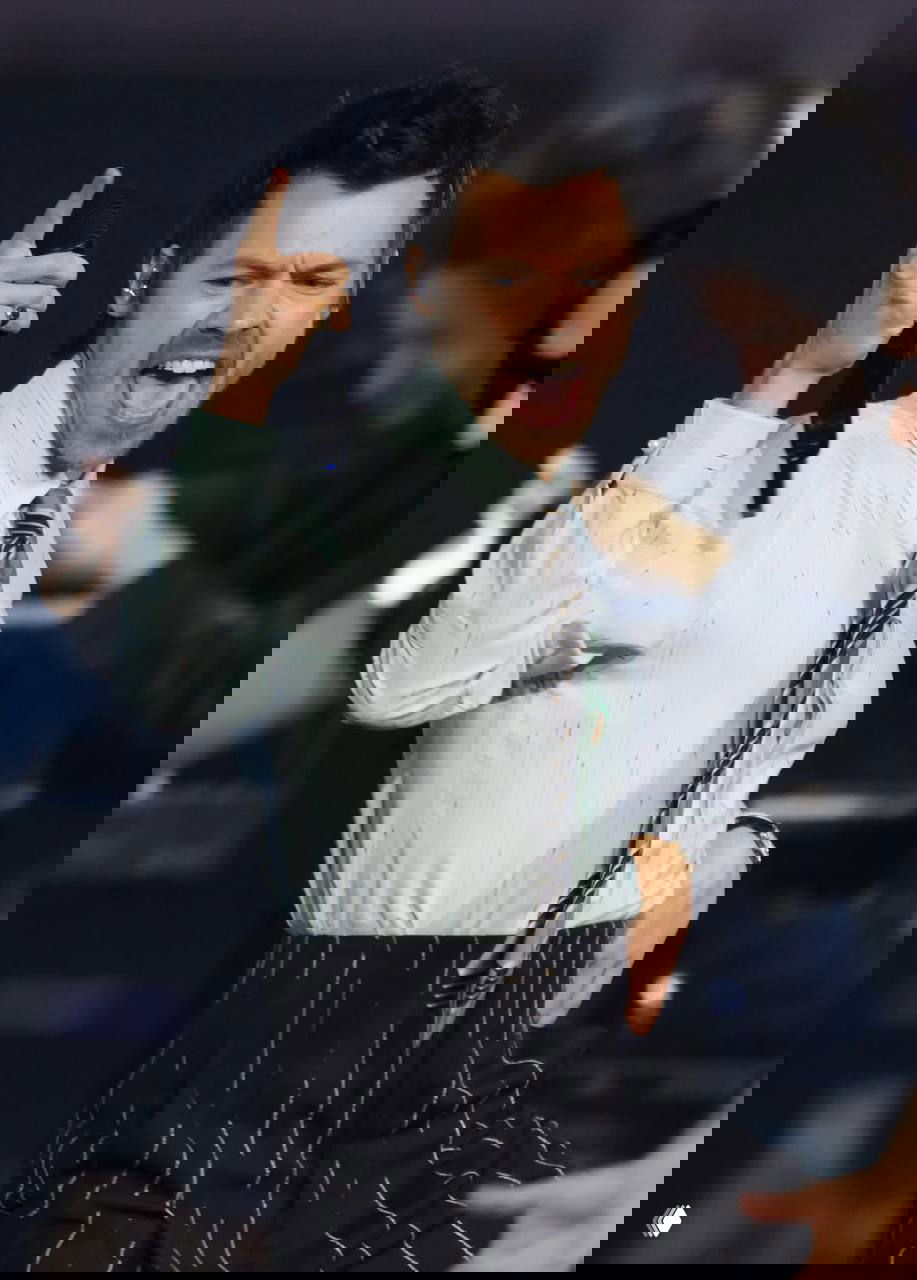 Крупный план исполнителя в рубашке и галстуке с микрофоном, эмоциональный момент выступления на сцене и бэк‑танцоры.