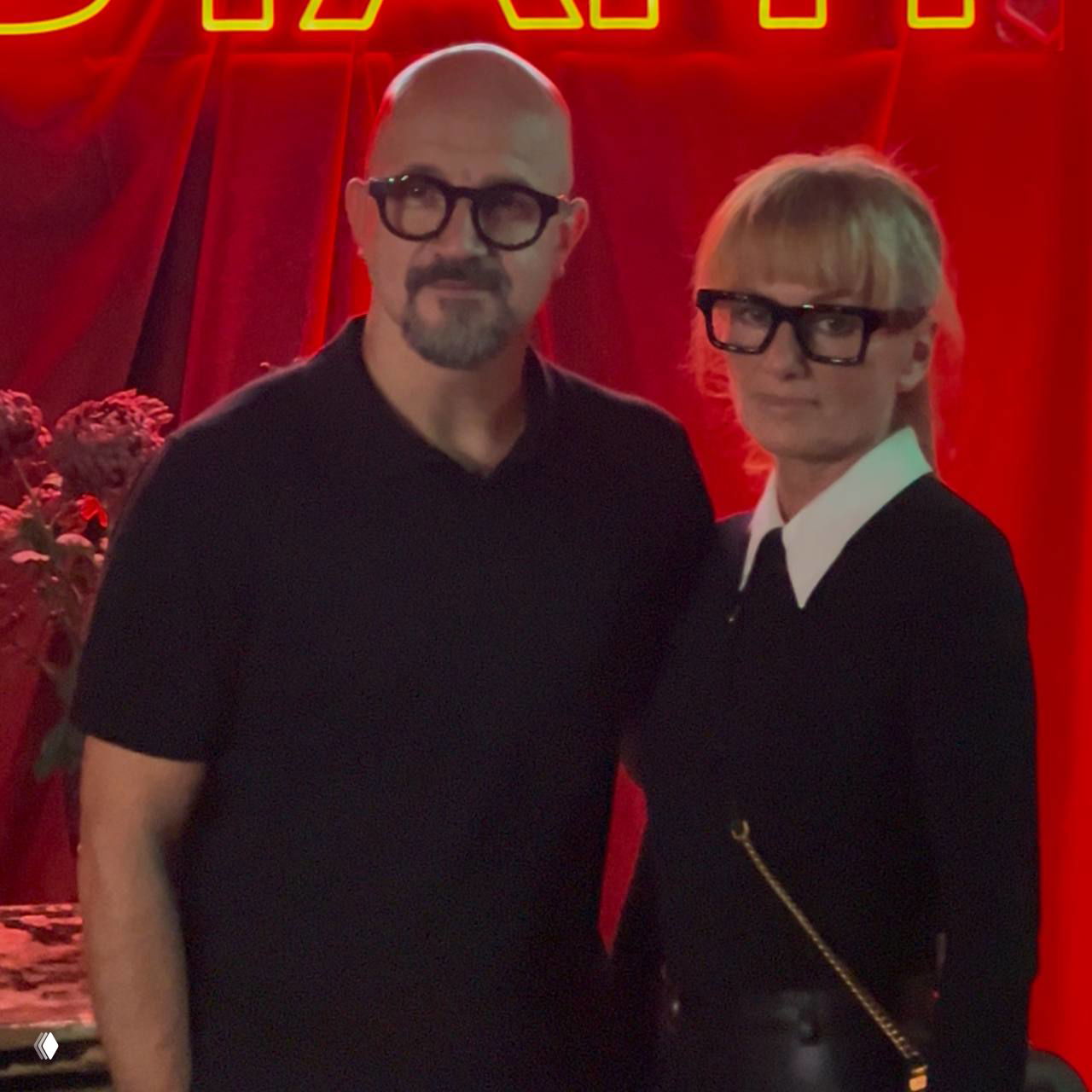 Пара гостей в очках позирует на фоне сцены презентации, вокруг видна красная подсветка и элементы оформления мероприятия.