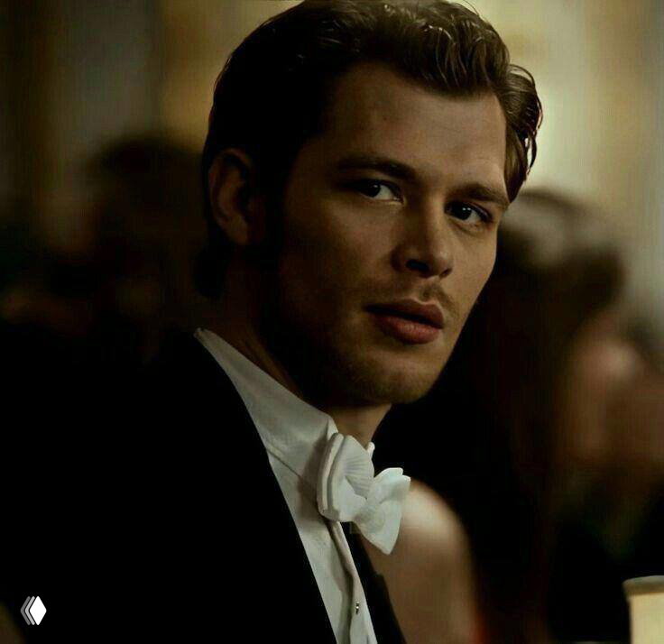 Элегантный мужчина в официальном костюме и бабочке, взгляд сдержан, образ тёмного аристократа с внутренними ранами и контролем.