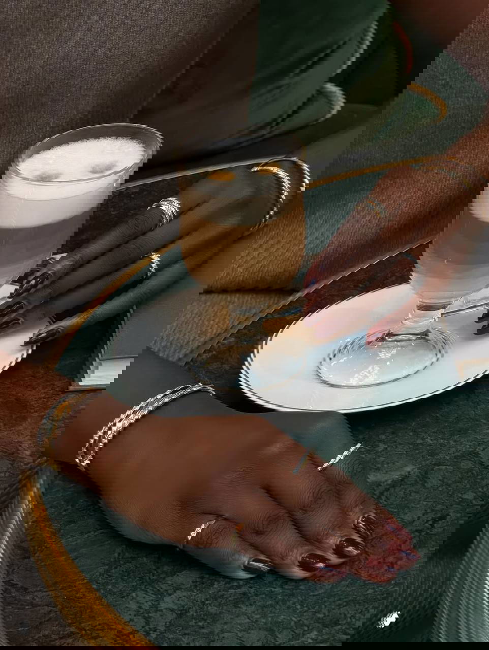 Руки с тёмно-бордовым классическим маникюром, украшенные золотыми кольцами; рядом латте на зелёном столе — элегантный образ.