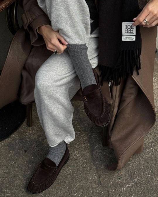 Фото в кадре: темно-коричневые лоферы из замши на ногах, комбинированные со светло-серыми трениками и тёмным пальто.