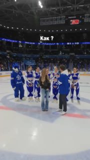 На льду хоккейной арены группа хоккеистов в форме окружает девушку, момент поздравления и помолвки, видно табло и трибуны.