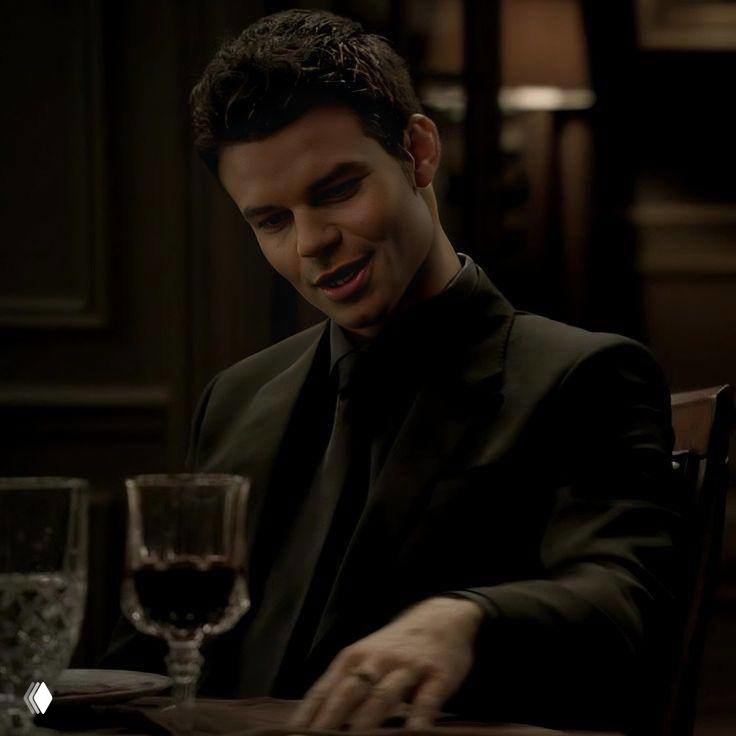 Мужчина за столом в тёмной обстановке, улыбка и жесты показывают близость и преданность — образ романтика-друга.