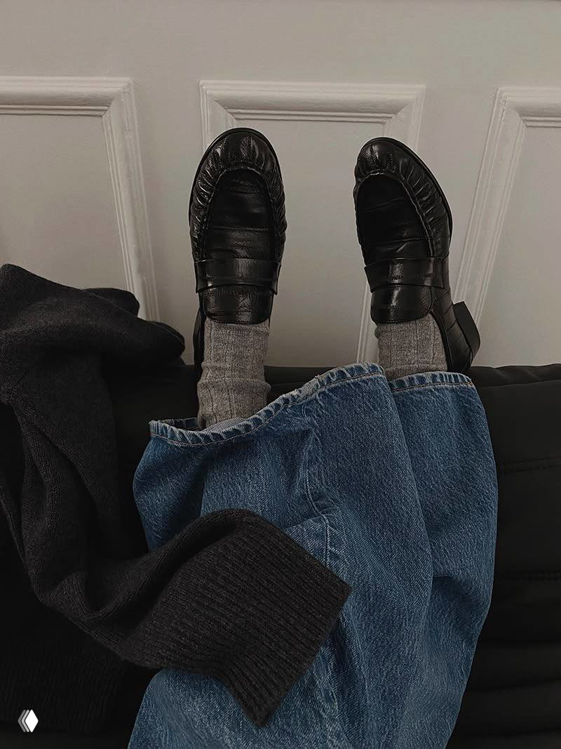 Черные глянцевые лоферы на ногах в серых носках, под широкие джинсы на фоне светлой декоративной панели — минималистичный уличный стиль.