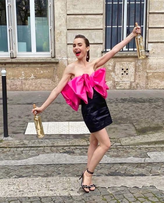Модель на улице в ярком розовом платье с объемным бантом на лифе, позирует с бутылками шампанского — уличная сценка в стиле модной съёмки.