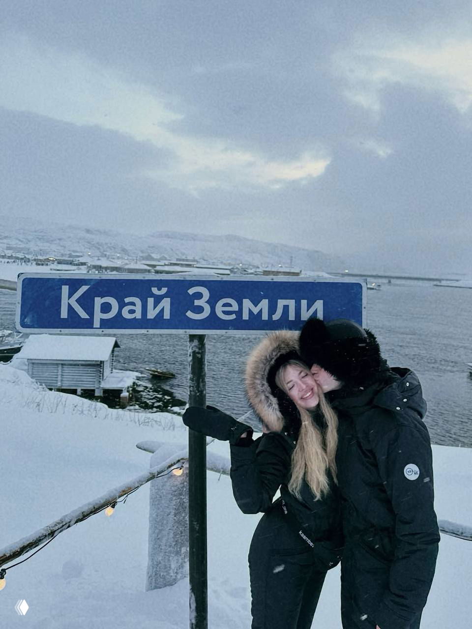 Аня и Артур у таблички «Край Земли» на побережье в Мурманске; пара обнимается и целуется на фоне заснеженного берега и воды