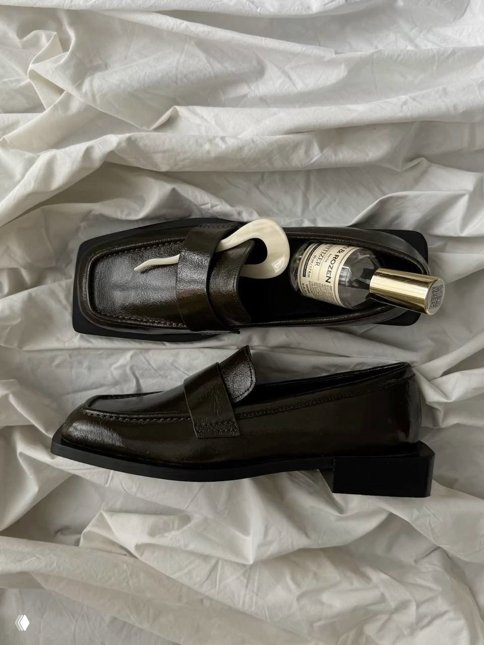 Плоская композиция: пара лакированных черных лоферов на светлой простыне рядом с флаконом парфюма — стилизованная товарная фотография.