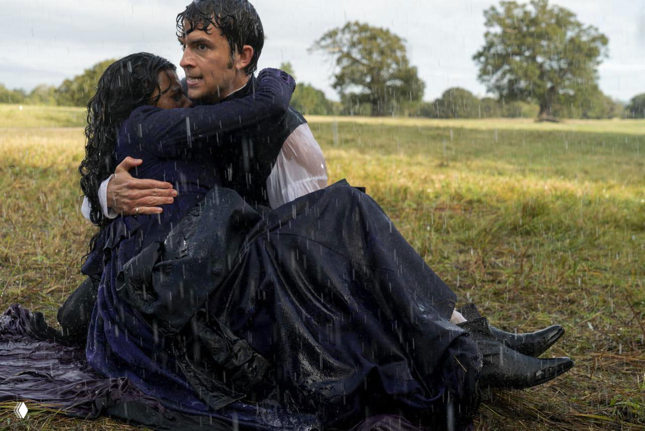 Драматический кадр на открытой местности под дождём: мужчина держит женщину на руках, одежда промокла — эмоциональная сцена на лужайке с мрачной погодой и напряжением.