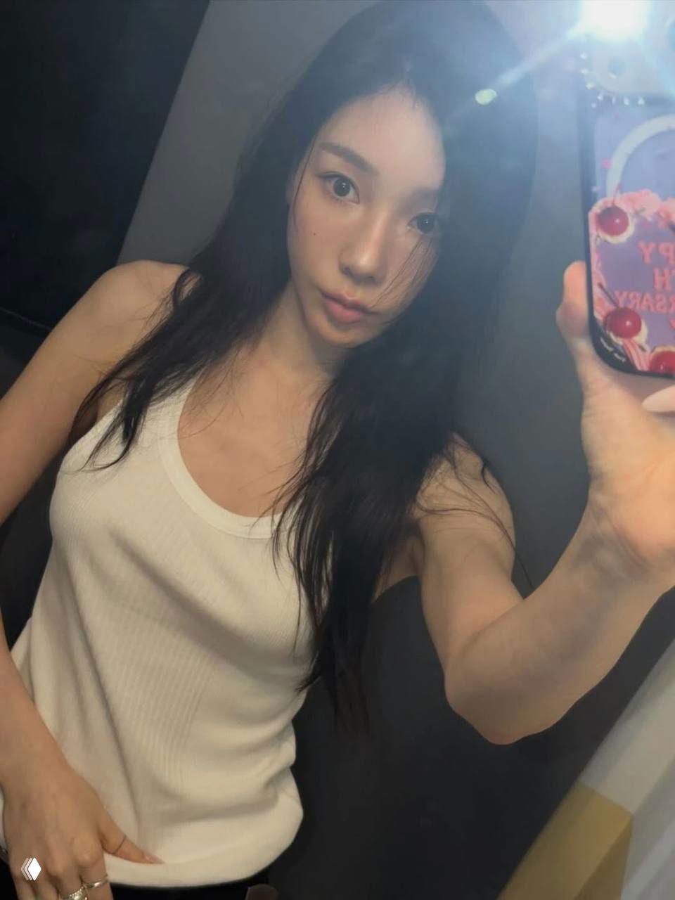 Фото в примерочной: девушка в белой майке делает селфи; видно тонкую стрелку в макияже и лёгкий контур лица при искусственном освещении.