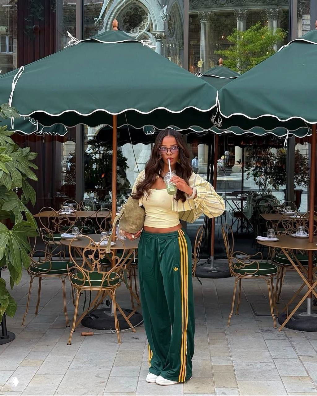 Девушка в зелёных спортивных брюках с жёлтыми лампасами на фоне уличного кафе под зелёными зонтиками, держит смузи
