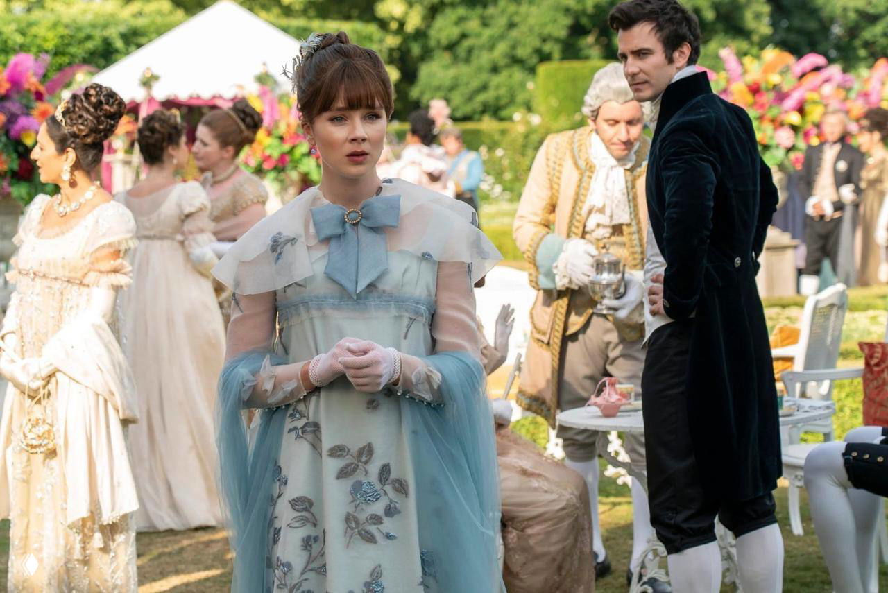 Дневная сцена в саду: женщина в нежно-голубом платье стоит на переднем плане, за ней мужчина в тёмном костюме — праздничная уличная обстановка с гостями и цветами.