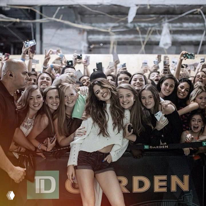 Фотография у сцены: женщина в центре кадра среди толпы фанатов, люди с телефонами и улыбками, атмосфера встречи и автографов.