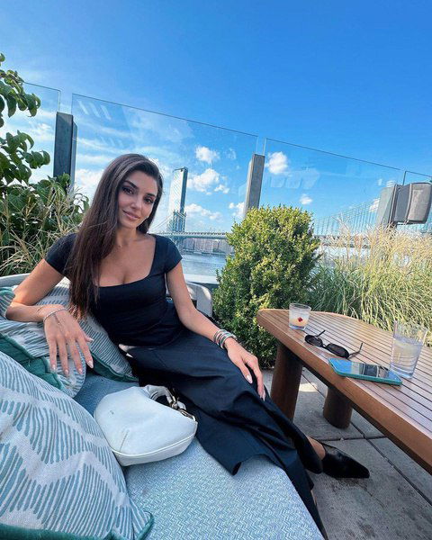 Женщина в чёрном повседневном платье сидит на открытой террасе у столика с городским видом и зеленью, расслабленный дневной образ на фоне города.