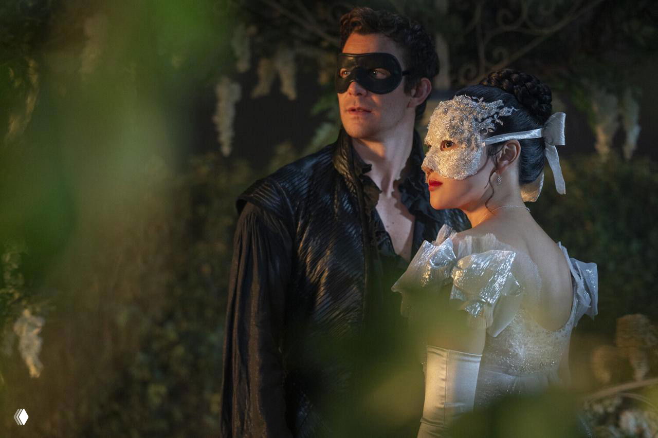 Ночная маскарадная сцена: пара в масках и вечерних одеждах стоит среди затемнённой зелени и подсветки — таинственный и атмосферный кадр бала.