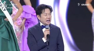 Ведущий или директор конкурса выступает на сцене «Мисс Вселенная», держит микрофон на фоне сценического оформления и конкурсанток.