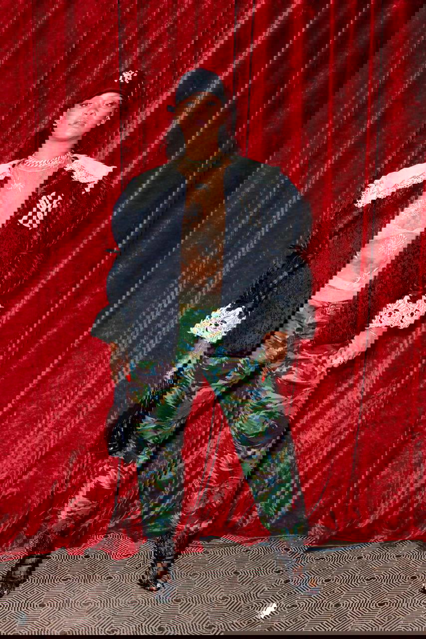 Фотография в помещении: пышный чёрный жакет с кружевной отделкой, камуфляжные брюки и бейсболка на фоне бархатной красной занавеси, вечерний образ.