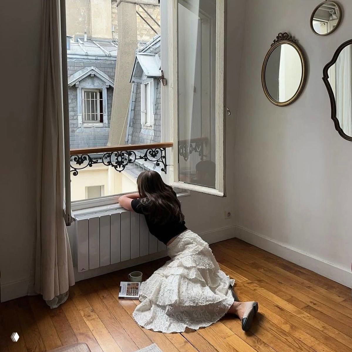 Девушка в длинной белой юбке у открытого окна с видом на старинные крыши, книга и чашка рядом — тихая моментная сцена.