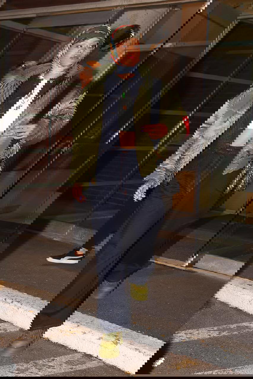 Утеплённая оливково‑зелёная бомбер‑куртка в сочетании с широкими тёмными джинсами и массивными зелёными ботинками — практичный городской образ при выходе из здания.