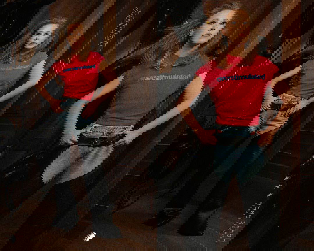 Фотография в полный рост: модель в джинсово-кожаных брюках, яркая красная футболка и уверенная поза на фоне снаряжения — рекламный сет IRNBY.