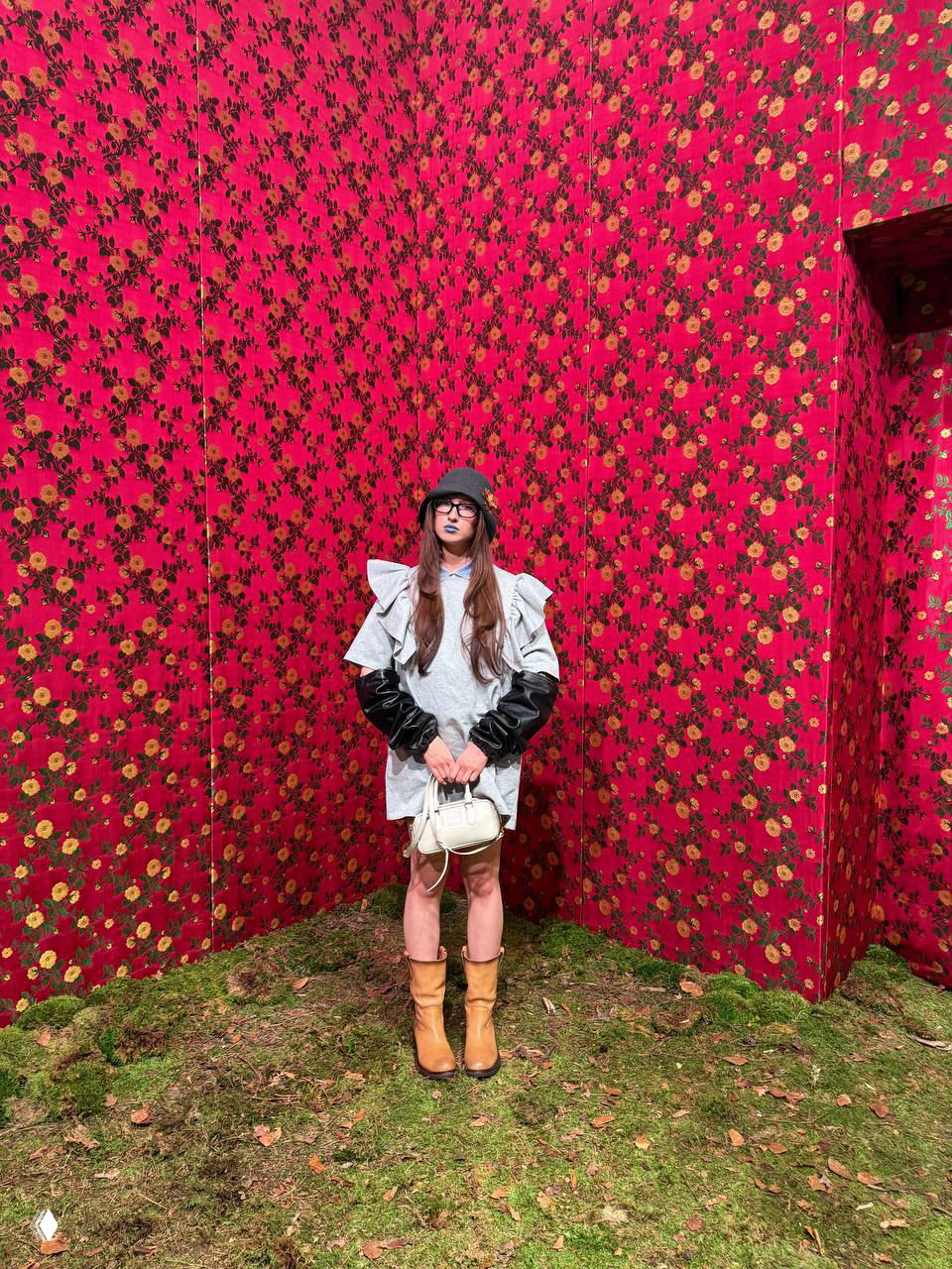 Полноформатный кадр: женщина в сером платье, коричневых сапогах и небольшой белой сумкой позирует на фоне стен с ярким цветочным принтом.
