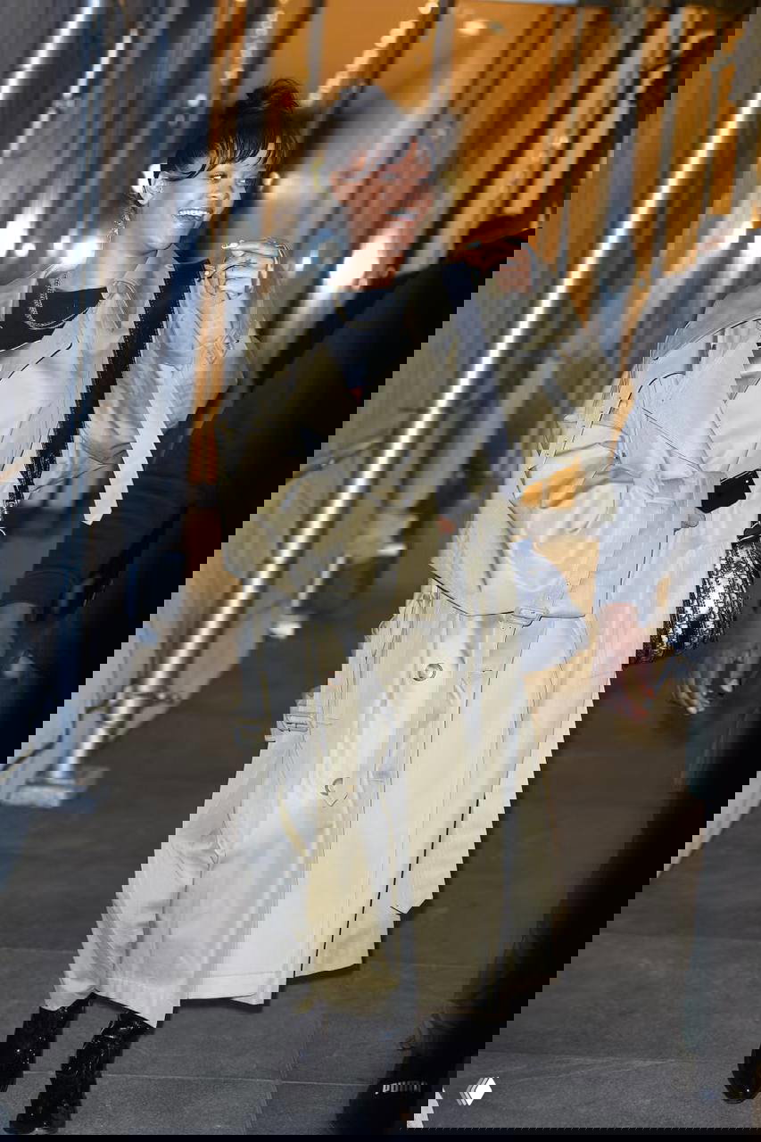 Ночное уличное фото: длинный песочный тренч, сумка через плечо и лакированные ботинки на освещённой городской улице, лёгкий смайлик и живое настроение.