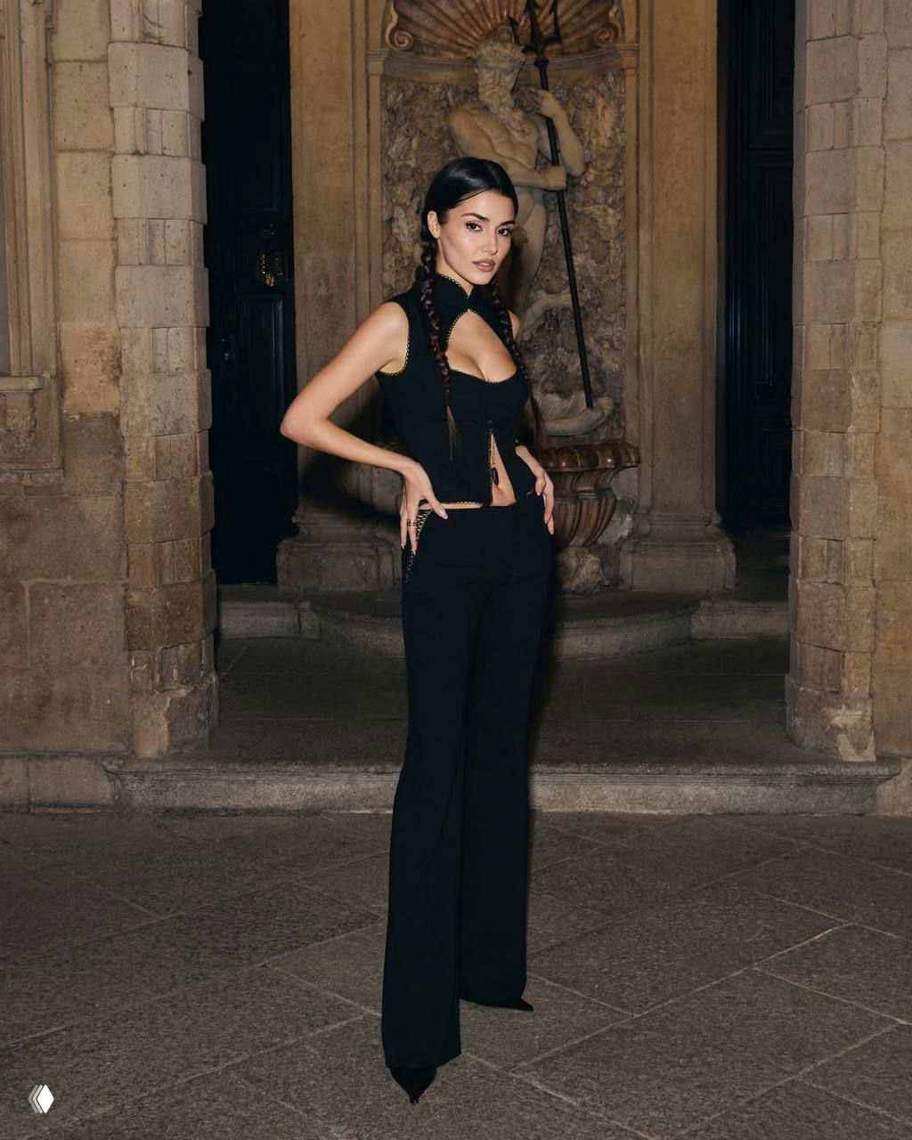 Ханде Эрчел у фонтана-нишки: полный рост в чёрном ансамбле, архитектурный фон и вечернее освещение.