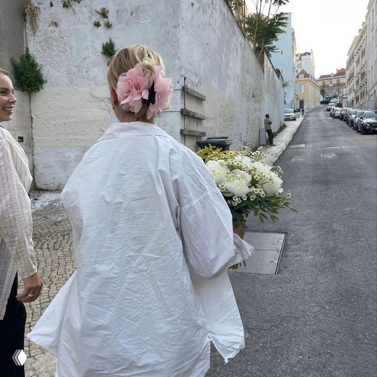 Девушка с цветком в волосах, идущая по узкой улочке с букетом белых цветов в руках — романтичный кадр в светлой, летней эстетике.