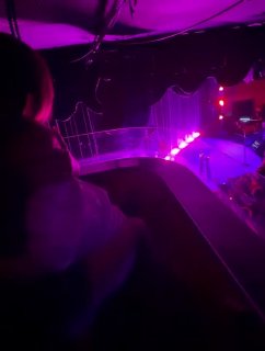 Тёмный кадр с пурпурным освещением сцены и силуэтом зрителя с балкона, передача концертной атмосферы.