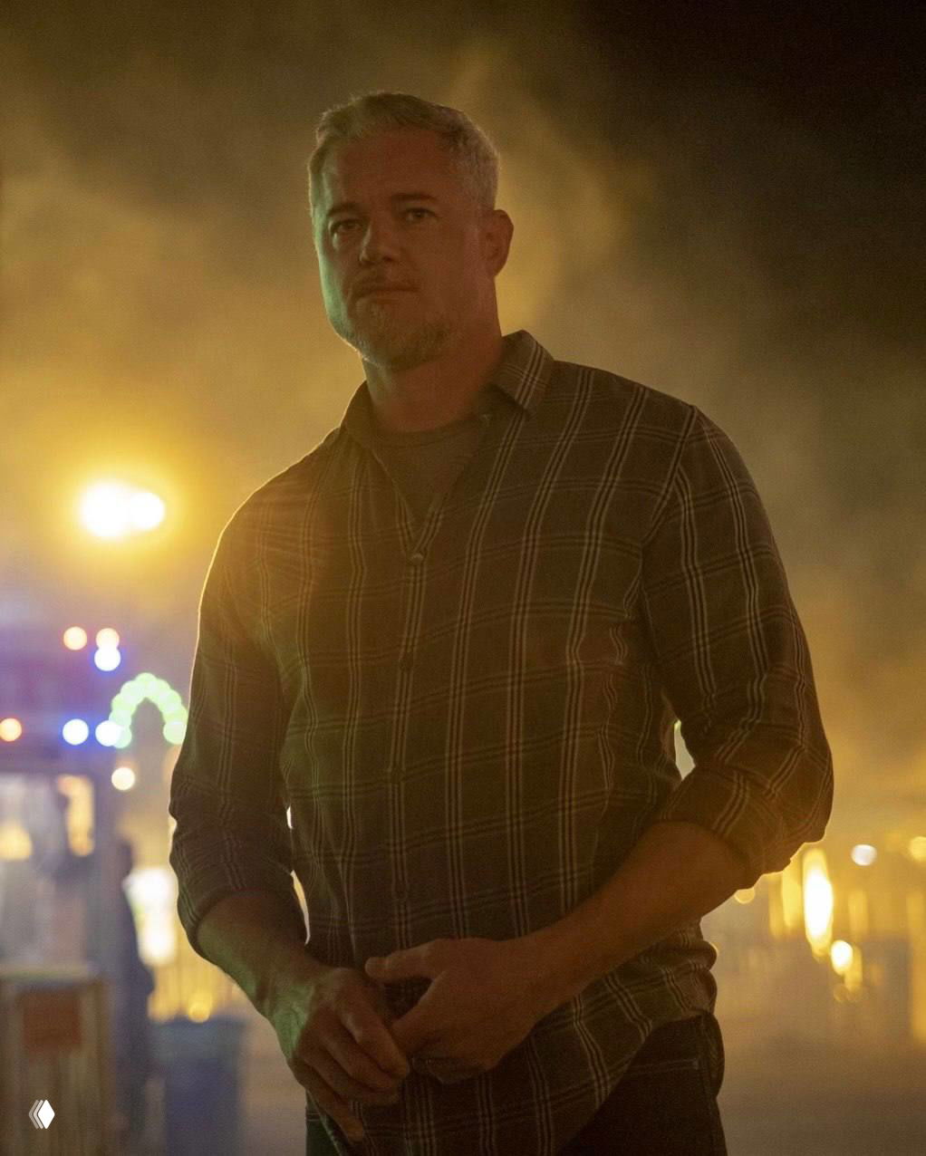 Мужчина в клетчатой рубашке на ночной площадке с тёплой подсветкой и дымом на заднем плане, кадр в драматичном освещении.