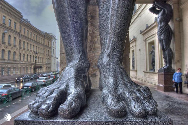 Крупный план каменных ног атлантов у фасада в Петербурге: детальная фактура скульптуры, мокрый камень и уличная сцена на заднем плане.