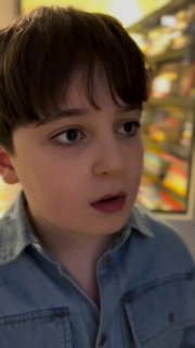 Крупный план задумчивого мальчика в магазине на фоне полок с товарами; выражение лица удивлённое, мягкое освещение.