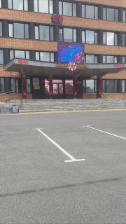 Фасад здания с центральным входом, лестницей и пустой парковочной зоной; над входом крупный экран — иллюстрация локации для выездов и встреч.