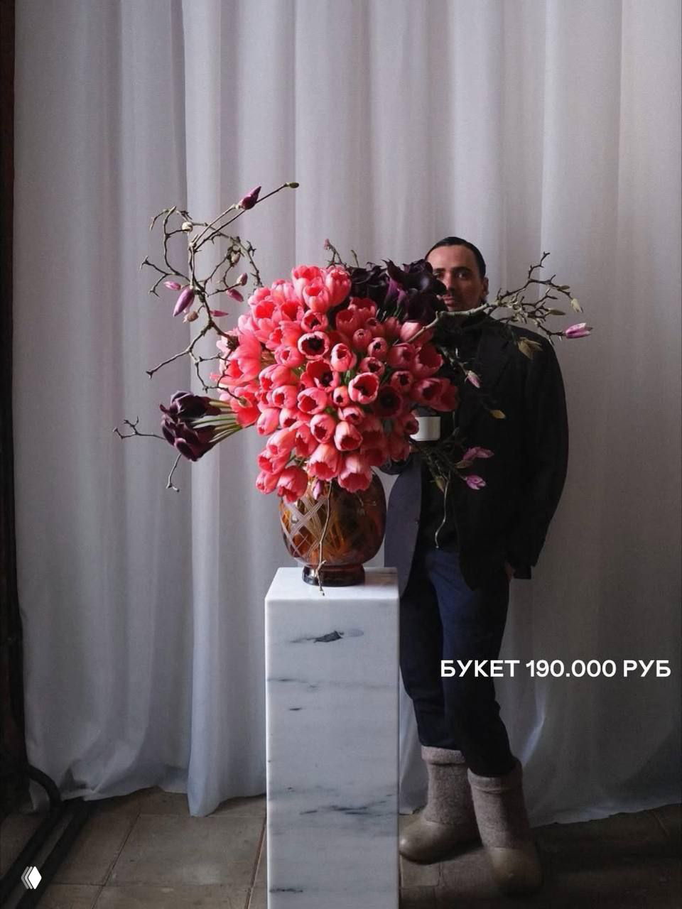 Большая композиция тюльпанов в вазе рядом с мужчиной в валенках на фоне тех же штор, контраст цены и повседневности видно на фото.